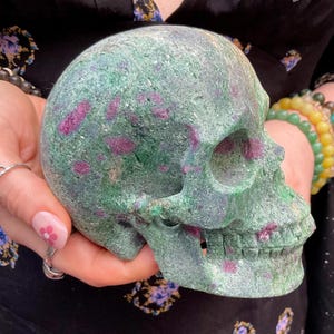 Puede incluir: Una escultura de calavera tallada en piedra verde con inclusiones rojas. La calavera se sostiene en las manos y presenta rasgos faciales detallados, incluyendo cuencas de los ojos, cavidad nasal y dientes. La piedra tiene una superficie texturizada.