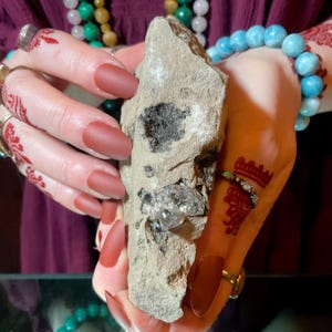 May include: A beige rock with embedded crystals and dark inclusions is held between two hands. The hands have long, matte red nails and are adorned with rings, bracelets, and henna tattoos. The background is a dark, blurred maroon.