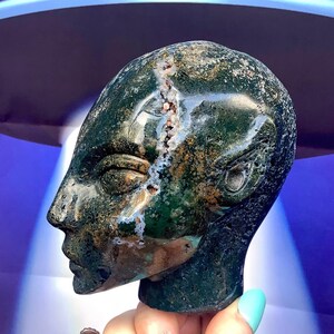 May include: A carved stone head sculpture in shades of green, brown, and white. The sculpture has a detailed profile with a visible crack running down the center. The head is held by a hand with a turquoise nail polish.