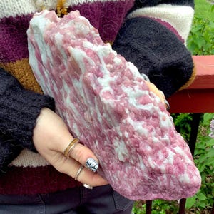 May include: A large, raw mineral specimen featuring a rough, crystalline texture with a prominent color palette of vibrant magenta-pink and opaque white. The stone displays jagged, uneven edges and a dense, layered structure throughout its surface.