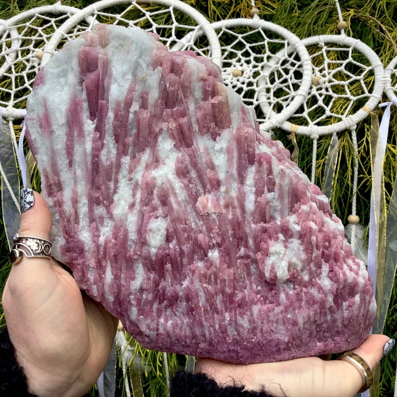 May include: A large, raw mineral specimen featuring vertical, elongated pink tourmaline crystals embedded within a white quartz matrix. The cluster displays a rough, natural texture with varying shades of magenta and deep rose throughout the stone.