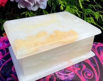 May include: A rectangular box crafted from polished white stone featuring translucent qualities and natural golden-yellow veining across the lid and sides. The box has a smooth, flat surface and sits on a matching rectangular stone base.