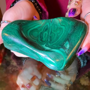 May include: A polished malachite stone featuring deep forest green and lighter emerald green concentric banding patterns. The stone has a smooth, rounded, and irregular shape with a glossy finish that reflects light across its surface.