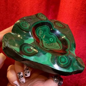 May include: A polished malachite bowl featuring deep green hues and distinct concentric circular banding patterns. The surface is smooth and glossy, showcasing natural mineral layers in various shades of dark and vibrant green throughout the stone.