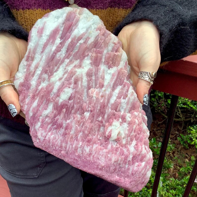May include: A large, irregular slab of raw pink tourmaline crystal embedded in a white quartz matrix. The specimen features elongated, striated pink mineral formations that contrast with the opaque white mineral base throughout the surface.