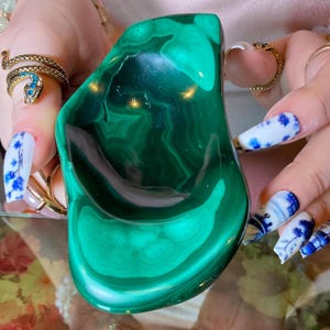 May include: A polished malachite bowl featuring deep forest green hues and characteristic concentric banding patterns. The stone has a smooth, glossy finish with a natural, irregular organic shape that highlights the intricate mineral layers.
