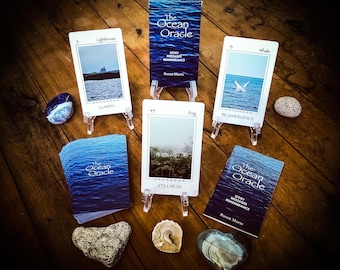 May include: An oracle card deck titled "The Ocean Oracle" with cards displayed on clear stands. The cards feature ocean-themed imagery and text, including "Lighthouse," "Whale," and "Fog." Additional items include stones and a seashell.