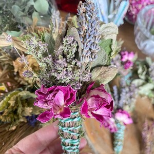 May include: A bundle of dried herbs and flowers featuring vibrant magenta rose heads, light purple lavender sprigs, and green foliage. The stems are tightly bound together with green and white striped twine, creating a compact floral arrangement.