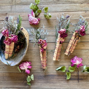 May include: Five bundles of dried herbs and flowers tied together with twine. The bundles are made with sage, lavender, and rose petals. The bundles are arranged on a wooden surface.