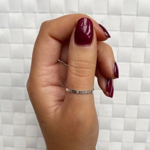 May include: A silver ring with a thin band and an inscription on the top. The ring is worn on the thumb of a hand with burgundy nail polish.
