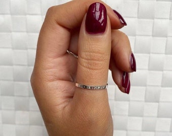 May include: A silver ring with a thin band and an inscription on the top. The ring is worn on the thumb of a hand with burgundy nail polish.
