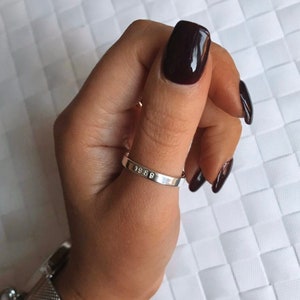 May include: A silver ring with the inscription "1989" on the band. The ring is worn on the thumb of a hand with dark burgundy nail polish.