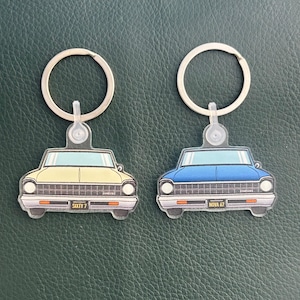 May include: Two car-shaped keychains. One is yellow with the text "SIXTY 7" on the license plate, and the other is blue with "NOVA 67". Both have silver key rings and clear plastic attachments.
