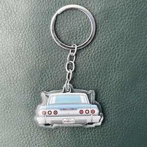Op de afbeelding: Een zilverkleurige metalen sleutelhanger met een doorzichtige acryl hanger in de vorm van de achterkant van een lichtblauwe oldtimer. De auto heeft rode ronde achterlichten, een chromen bumper en een kentekenplaat met de tekst MENCHACA.