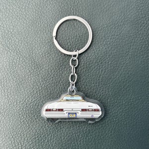 May include: A silver metal keychain featuring a white convertible car charm. The car is depicted from the rear view with red taillights, a tan interior, and a blue license plate that clearly displays the text CLB in yellow lettering.