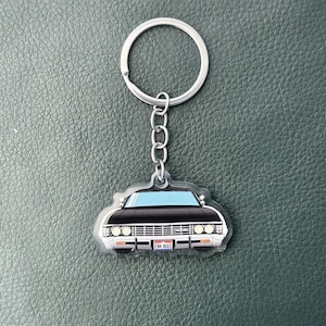 May include: A keyring featuring a flat, acrylic charm shaped like the front view of a black vintage car. The car has a light blue windscreen, round headlights, and a number plate that reads CNK 8003. It is attached to a silver metal ring.