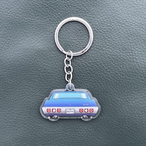 May include: A keychain featuring a blue car rear view graphic with red taillights, a silver bumper, and a license plate displaying the text 0003783. The charm is attached to a circular silver metal split ring by a short silver chain.