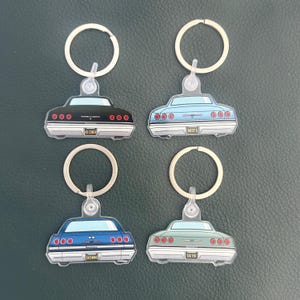 Op de afbeelding: Vier sleutelhangers met het achteraanzicht van klassieke auto's in zwart, lichtblauw, donkerblauw en saliegroen. Elke sleutelhanger heeft een zilveren metalen ring en een nummerplaat met 65 CHEV, MIST 5, SIXSWAG of SIX FIV.