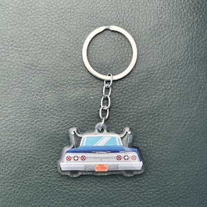 May include: A keychain featuring a flat, illustrated charm of the rear view of a dark blue vintage car. The car has a chrome bumper, red circular taillights, and an orange license plate with the text MSH5. It includes a silver metal ring.