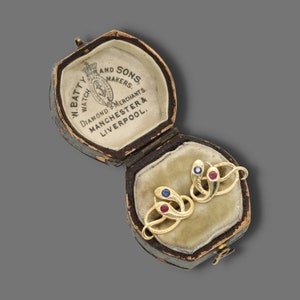Peut inclure: Une petite boîte en bois avec un couvercle à charnière. Le couvercle est décoré d'une inscription dorée qui dit "W. Batty & Sons, Watch Makers, Diamond Merchants, Manchester & Liverpool". À l'intérieur de la boîte se trouvent deux broches en forme de serpent en or avec des pierres précieuses rouges et bleues.