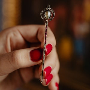 Puede incluir: Broche plateado en forma de llave adornada. La parte superior presenta una perla y filigranas decorativas. El tallo está adornado con pequeñas piedras rojas. Una joya clásica y elegante.