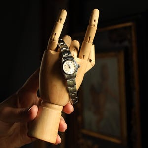 Può includere: Un orologio da polso argentato con quadrante chiaro è esposto su una mano di legno che fa il gesto del rock. L'orologio ha un cinturino in metallo ed è tenuto su uno sfondo scuro, con opere d'arte incorniciate visibili.