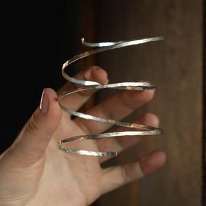 Puede incluir: Un brazalete de plata con textura martillada, que se enrolla en espiral alrededor del brazo. El brazalete se sostiene en una mano, mostrando su diseño. La pieza de joyería tiene una estética minimalista y elegante.
