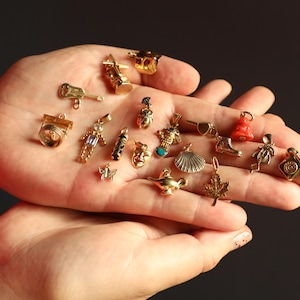 May include: A selection of small, gold-coloured charms in diverse shapes, such as a guitar, a boot, a seashell, and a genie's lamp. The charms are held in an open hand against a dark backdrop.
