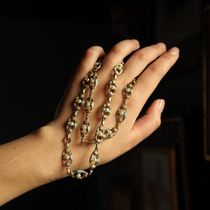 May include: Gold-toned necklace with an intricate design featuring oval links, small pearls, and turquoise-colored accents. The necklace is held in a hand against a dark background, highlighting its delicate details and vintage aesthetic.