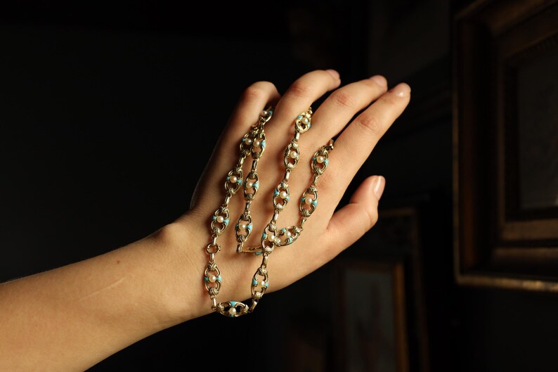 May include: Gold-toned necklace with an intricate design featuring oval links, small pearls, and turquoise-colored accents. The necklace is held in a hand against a dark background, highlighting its delicate details and vintage aesthetic.