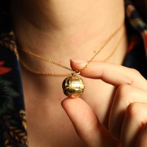 May include: A gold-colored necklace with a spherical pendant. The pendant has a textured surface and hangs from a delicate chain. The necklace is worn against a dark floral patterned background.