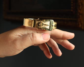 May include: A gold-colored bracelet with a buckle design. The bracelet is held on a hand, showcasing its metallic sheen and intricate details. The buckle is rectangular, and the bracelet appears to be made of a solid metal.