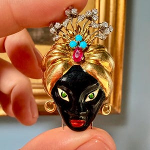 Op de afbeelding: Een vintage broche met een zwart gezicht met rode lippen en groene ogen, versierd met een gouden tulband. De broche is versierd met turkooizen stenen, een robijn en diamantaccenten, vastgehouden door een hand.