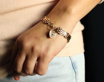 May include: A gold-toned bracelet with a heart-shaped charm. The bracelet features a series of rectangular links and a small heart-shaped charm with a keyhole design. The bracelet is worn on a wrist.