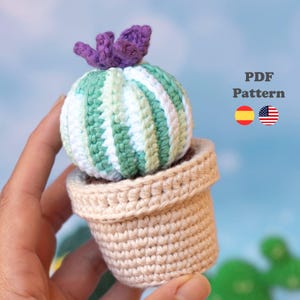 Puede incluir: Un cactus de ganchillo con rayas verticales en verde claro, verde oscuro y blanco, rematado con una flor morada, en una maceta de ganchillo beige. La imagen incluye el texto PDF Pattern con iconos de las banderas de España y Estados Unidos.