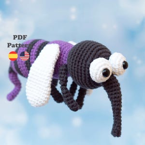 Puede incluir: Un juguete de mosquito tejido a ganchillo con cabeza negra, una probóscide larga y negra, y grandes ojos blancos con pupilas negras. El cuerpo tiene rayas moradas y negras con alas blancas. El texto dice PDF Pattern con iconos de banderas.
