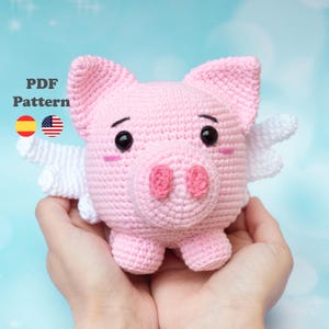 Puede incluir: Un cerdo rosa tejido a mano con alas blancas, ojos negros y un hocico rosa. El cerdo se sostiene con dos manos. El texto "PDF Pattern" está en la esquina superior izquierda, con las banderas española y estadounidense debajo.