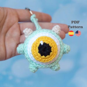 May include: A round, crocheted keychain featuring a central eye design with a black pupil, a yellow iris, and a white sclera. The outer body is light mint green with four small, protruding appendages and a matching crocheted loop at the top.