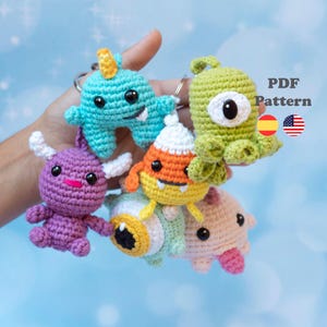May include: A collection of six small crocheted monster keychains in various colors including teal, purple, green, orange, and pink. The creatures feature plastic safety eyes, small horns, and appendages. Text reads PDF Pattern with Spanish and US flags.