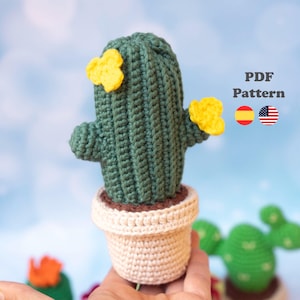 May include: A dark green crocheted cactus with two yellow flowers, planted in a beige crocheted pot. The image includes the text "PDF Pattern" along with Spanish and American flag icons, indicating a digital pattern for this crocheted item.