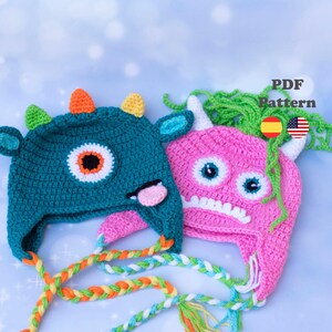 Puede incluir: Dos gorros de monstruo de ganchillo. Uno es verde azulado con cuernos naranjas y amarillos, un ojo blanco y negro y una lengua rosa. El otro es rosa con cuernos blancos, dos ojos azules y una boca blanca. Ambos tienen lazos trenzados.