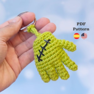 Puede incluir: Un llavero de mano de ganchillo color verde lima con detalles de costura negros en la palma. El artículo incluye una anilla metálica y el texto PDF Pattern con iconos de banderas de España y EE. UU. que indican los idiomas.