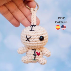 Puede incluir: Un llavero de muñeco vudú tejido a ganchillo en color crema con un ojo de plástico negro grande, un ojo en forma de X bordada y un pequeño detalle de corazón rojo en el pecho. El texto PDF Pattern aparece junto a iconos de banderas.