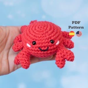 Puede incluir: Un cangrejo rojo tejido a ganchillo con ojos negros y una boca sonriente. El cangrejo tiene pequeños acentos blancos en las mejillas y se sostiene contra un fondo de cielo azul. Incluye el texto "PDF Pattern" y banderas.
