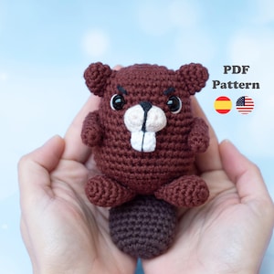 Pode incluir: Um castor marrom de crochê com dentes brancos e olhos pretos. O castor é segurado em duas mãos. A imagem inclui o texto "PDF Pattern" e bandeiras da Espanha e dos Estados Unidos.