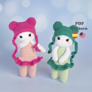 May include: Two crocheted dolls wearing pink and green dresses and hats. The dolls have white faces with black eyes and are standing on a light blue background. PDF Pattern.