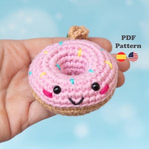 Puede incluir: Un donut rosa tejido a ganchillo con chispas de colores y una cara sonriente. El donut tiene una base marrón y un pequeño trozo tejido a ganchillo de color beige en la parte superior. El texto "PDF Pattern" es visible.