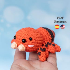 Puede incluir: Un juguete de araña tejido a ganchillo en color naranja y negro con grandes ojos negros y pedipalpos de color beige claro. El texto PDF Pattern es visible junto a un icono de bandera española y estadounidense. Cuerpo redondeado con rayas.