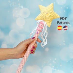 Puede incluir: Una varita mágica de ganchillo con una estrella amarilla y rosa. La estrella está adornada con pequeñas perlas. La varita tiene un mango rosa con flecos blancos y un acento de estrella púrpura. Incluye el texto "PDF Pattern".
