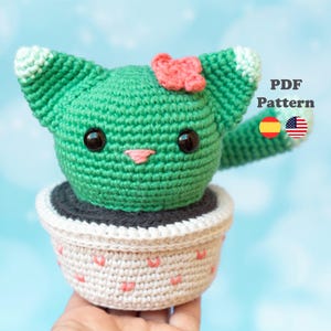 Puede incluir: Un gato cactus tejido a ganchillo en una maceta. El gato cactus verde tiene ojos negros, una nariz rosa y un pequeño lazo rosa. La maceta es blanca con detalles de corazones rosas. El texto "PDF Pattern" es visible.